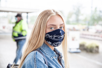 Eine jung, blonde, langhaarige Frau mit einem Mund-Nasenschutz gegen Covid19 steht am Bahngleis und...
