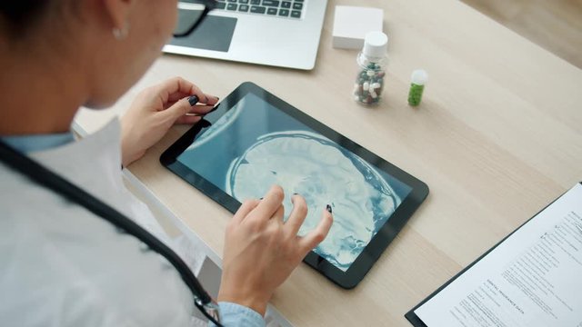Young woman doctor is studying MRI images on tablet screen working in office alone concentrated on patient's examination results. People and profession concept.