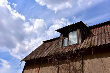 Old destroyed roof window with cracked wooden frame on top of ruins of abandoned suburban family house on the background blue sky with white clouds. An attic room in the roof of a house