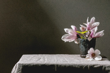Bouquet of beautiful pink magnolia flowers on branches with new leaves in ceramic vase, standing on gray linen table cloth. Copy space
