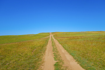 Fototapeta na wymiar The road to the field goes to the top of the hill. Bright sunny day and cloudless blue sky.