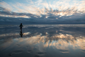 Silhouette of boy in the distance running out on the beach towards the ocean, stormy sky reflection