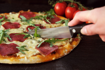 homemade pizza on a dark rustic background, next to a branch of tomatoes and arugula greens. A slice of pizza is cut off with a knife