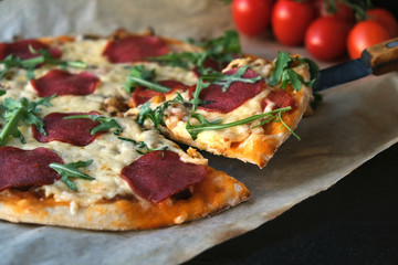 homemade pizza on a dark rustic background on craft paper, next to a branch of tomatoes and arugula greens. A slice of pizza is cut off with a knife