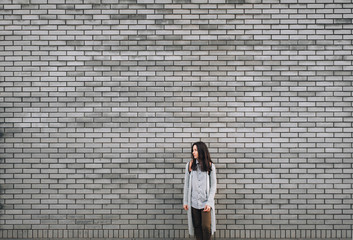 An young caucasian woman with a backpack standing half a turn near the gray brick wall. The pensive look of a lonely girl is directed to his right. Stone texture with copy space.