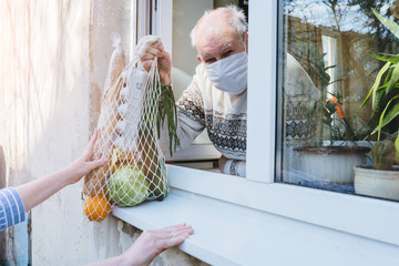 A sad elderly wrinkled woman is looking through the window in the protective mask of her house....