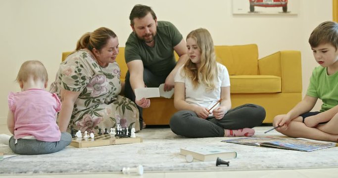 Family resting on floor and sofa at home. Chubby parents examining family pictures and showing photos to studying children while sitting on floor and sofa near baby playing with chess pieces during