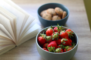 Bowl of chocolate chip cookies, bowl of strawberries and an open book on a table. Selective focus.