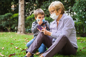Grandmother and little kid playing with a smartphone in the backyard while wearing protective masks...