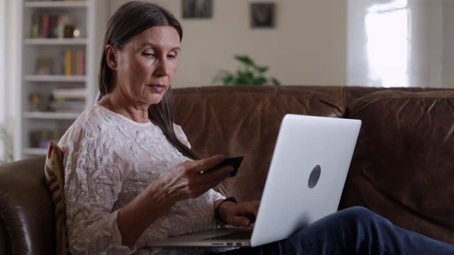Beautiful elderly lady is shopping in online store while sitting on sofa in apartment spbi. Elegant mature woman is making purchases in internet, using credit card and laptop on couch in home interior