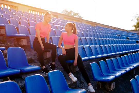 Two pretty sporty girls relaxing after workout outdoor. Two beautiful ladies relax after gym or running sitting on the seats at the stadium looking at each other smiling. Relax at sunset light.