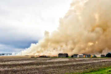 Obraz na płótnie Canvas Billowing clouds of smoke from a fire burning a farm field with old sheds nearby in a summer landscape