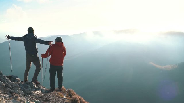 4K Two hikers on top of mountain holding hiking poles spreading arms around in fierce wind