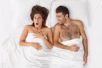 Obraz na płótnie Canvas Young newlywed couple wake up in morning. Frightened woman recalls something astonishing, cheerful husband lies near in comfortable bed under white sheet. People, home, relationship, bedding concept