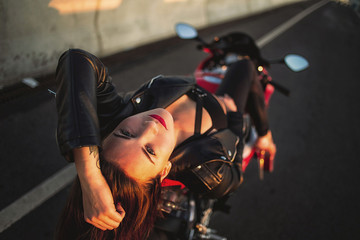 Fototapeta na wymiar Urban fashion. Outdoor portrait of pretty young woman in black leather jacket lying on red motorcycle on sunset