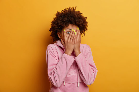 Intrigued curious dark skinned woman peeks through fingers, covers face with palms, hides herself, afraids of something, has bugged eyes, dressed in casual sweatshirt, poses indoor over yellow wall