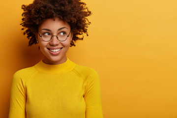 Pleased feminine woman looks with smile aside, has optimistic attitude, waits for good thing happen, concentrated mysteriously away on promo announcement, wears round glasses, yellow jumper.