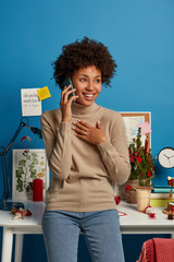 Cheerful curly haired female freelancer has discussion with coworker, enjoys telephone conversation, glad to hear something, feels joyful on remote job, stands in own cabinet near workplace.