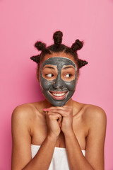 Happy beautiful woman takes good care of skin, applies facial mask for rejuvenation, keeps hands pressed toogether under chin, wears towel, has combed hair buns, looks aside. Pampering, wellness