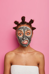 Dark skinned annoyed woman applies clay mask against acne on face, rejuvenates problematic skin, stands wrapped in bath towel, has hair buns, clenches teeth, isolated on pink background. Cosmetology