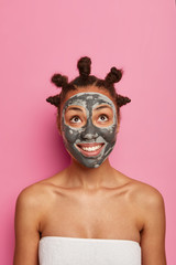 Facial cleaning, skin care and beauty treatments concept. Pleased dark skinned woman looks above with delight, applies clay nourishing mask for rejuvenation stands wrapped in towel after taking shower
