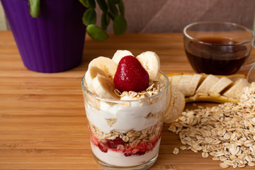 Granola with yogurt, banana and strawberries on a background of a cup of coffee and a flower in a pot