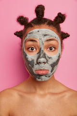 Close up portrait of sad mixed race young woman looks at herself in mirror, applies clay mask on face, stands with bare shoulders, cares about body and complexion, isolated over pink background.