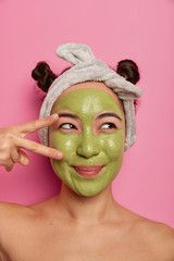 Pretty dreamy young female model makes peace gesture over eye, applies nourishing green mask on face, wears headband, stands naked indoor, enjoys beauty treatments, isolated on pink studio wall