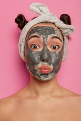 Funny mixed race young woman pouts lips, applies facial clay mask, looks directly at camera, has two combed buns, poses naked indoor, tries new beauty product, glad to have fresh clean skin.