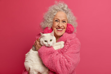 Obraz premium Portrait of wrinkled grandmother holds white cat on hands, stays at home during pandemic outbreak, wears fluffy earrings, soft robe, going to feed pet, isolated on pink background. Woman on pension