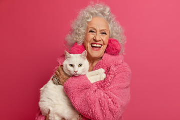 Portrait of aged wrinkled woman enjoys good day with favorite white cat, wears domestic robe, feels not bored on retirement, going to feed pet, loves animals, poses indoor against pink background