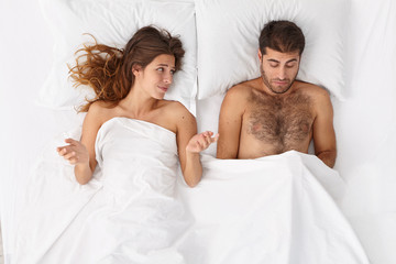Disappointed man has erectile dysfunction during sex, his woman partner lies near under white blanket, puzzled with husbands impotence, spreads hands sideways. Sexual problems. Men health concept