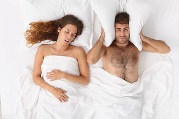 Frustrated man covers ears with pillow, cannot fall asleep because of loud wife snoring, suffers from insomnia, pose in bedroom. Tired man irritated by womans snoring. People, health, sleep disorder