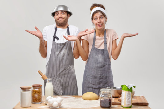 Hesitant Questioned Young Woman And Man Shrug Shoulders, Stand Together In Aprons, Dont Know What To Cook Or Which Ingredients Add, Make Dough For Pie. Bakery Workers At Kitchen Prepare Tasty Cake