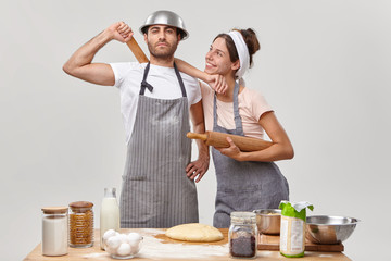 Proud man and his cheerful wife busy at kitchen, dressed in aprons, finish making dough, bake bread together, use secret ingredient, stand against white background near table with fresh products