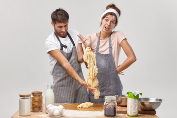 Clumsy man in apron stretches sticky dough, has difficulties during cooking, indignant housewife stands near, cannot understand whats wrong. Bad cookers at kitchen. Culinary courses for amateurs