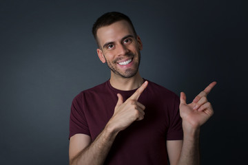 Advertisement concept. Positive, pretty, caucasian man with beaming smile in shirt on grey background is pointing with his two index finger and looking on empty copy space.