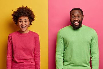 Shot of happy smiling Afro American couple express positive emotions, wear pink and green jumpers, enjoy pleasant moment, giggle over funny situation happend with them, pose over two colored wall - Powered by Adobe