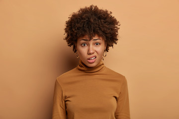 Fototapeta na wymiar Puzzled African American woman has displeased expression, frowns face, looks with disgust at something unpleasant, dressed in casual wear, isolated on brown background. Emotions and reaction