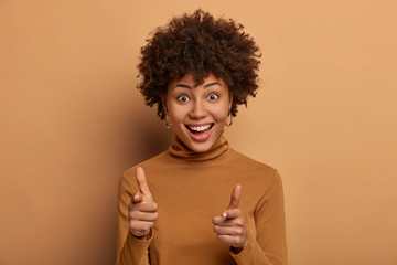 Check it out. Happy curly woman makes finger gun gesture, welcomes friend, chooses someone, has broad satisfied smile, dressed in casual turtleneck, isolated on beige background. You are what we need