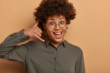 Fototapeta na wymiar Whos calling? Positive Afro American woman imitates phone call, makes mobile gesture and waits for answer, smiles broadly, dressed in formal shirt, asks colleague give telephone number to keep contact