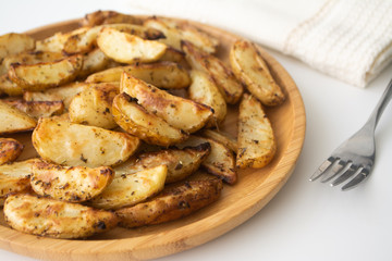 Oven-baked potato wedges with dried herbs on a wooden plate with a napkin and a fork in the background