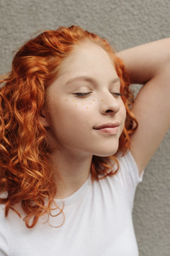 The portrait of the curly red hair girl with heart shaped fake freckles staying near the wall on the street with closed eyes