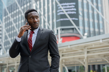 Young handsome African businessman talking on mobile phone in front of modern building at Bangkok city