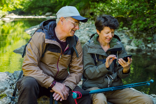 Smiling couple of travelers uses smartphone near mountain river. Summer vacations in mountains. - Powered by Adobe