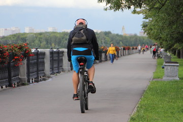 One man in sportswear and a bike helmet with headphones rides a Bicycle along the embankment alley on a summer day, a view from the back