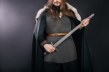 Knight in chain mail and with a fur collar on a black cloak on a gray background. Portrait of a Viking man with long hair and a beard in armor holds a sword. Studio photo