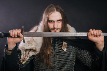Knight in chain mail and with a fur collar on a black cloak on a gray background. Portrait of a Viking man with long hair and a beard in armor holds a sword. Studio photo