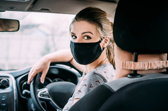 A young woman in a black medical mask and blond hair and a gray T-shirt is sitting at the wheel of a left-hand drive car. Portrait photo. Close up. Quarantine concept. Virus. Pandemic.