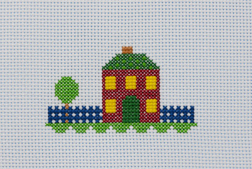 The cross-stitch colored house is in summer. The embroidered picture on the white canvas, close-up.